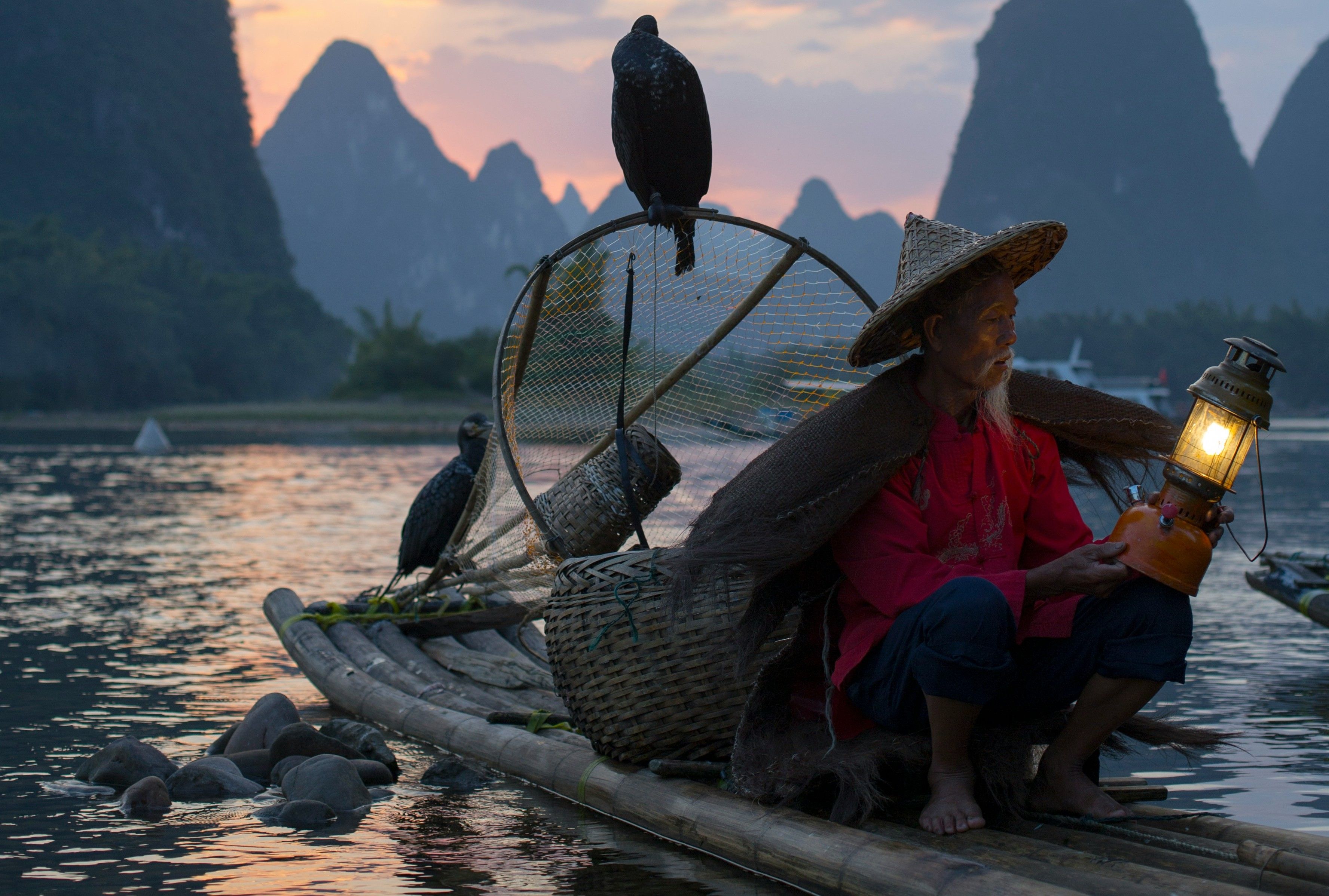 Un pêcheur traditionnel sur un radeau de bambou avec ses cormorans au coucher du soleil dans un paysage montagneux