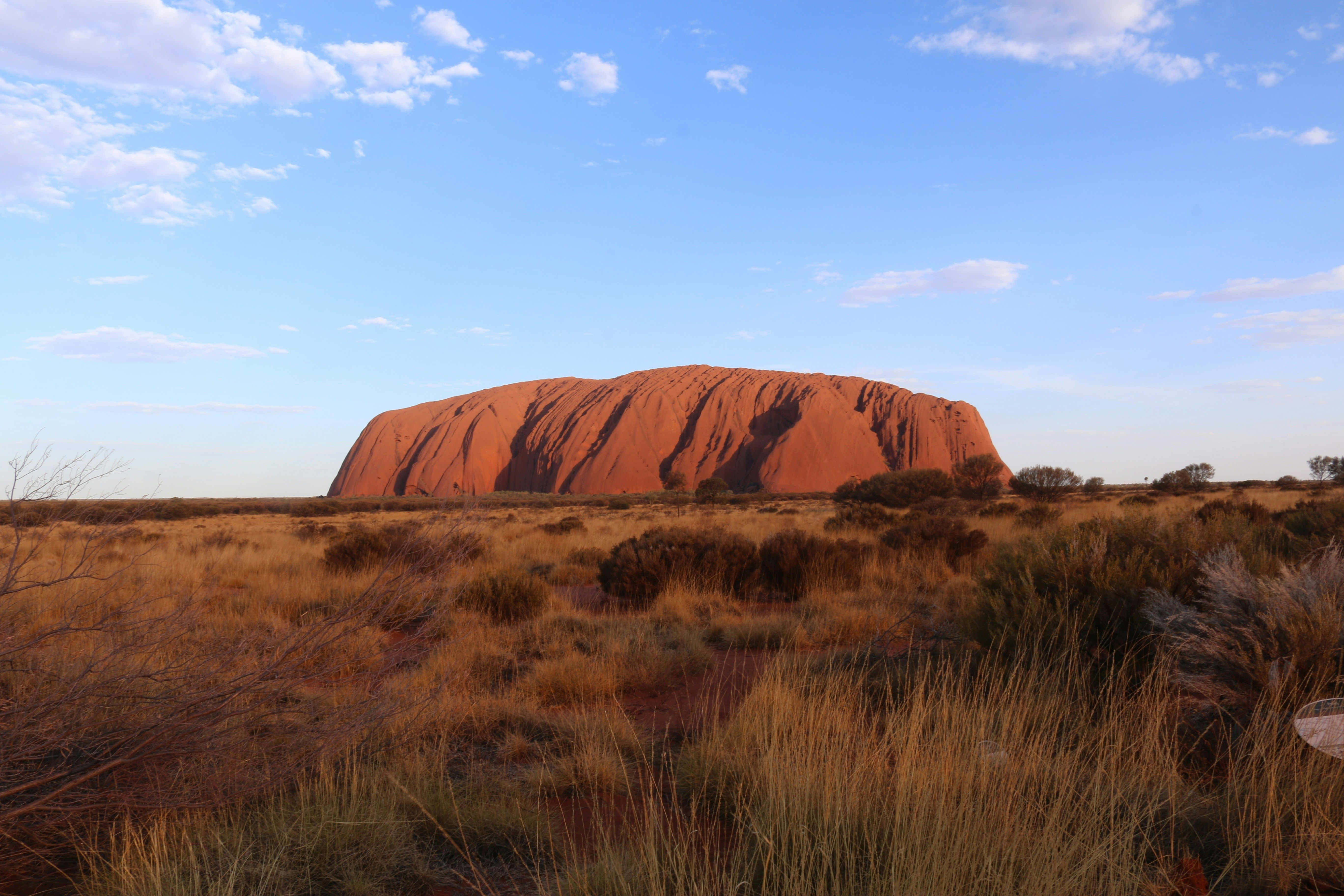 Voyage en Australie - Vaste monolithe d'Uluru baigné par la lumière du soleil couchant au milieu de la végétation sauvage du désert Vaste monolithe d'Uluru baigné par la lumière du soleil couchant au milieu de la végétation sauvage du désert